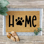 Dog paw print home doormat featuring a natural coir fiber texture with black lettering, layered over a gray and white geometric patterned rug on a wood floor, perfect for a pet lover entryway or front porch decor welcome mat.