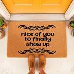 Funny welcome doormat with the text nice of you to finally show up, featuring a natural coir fiber texture and black decorative scroll accents, placed on a porch in front of a bright orange door for a humorous home entryway decor