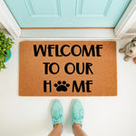 Dog paw print welcome doormat for pet lovers, featuring a natural coir fiber texture with black text that reads Welcome To Our Home, placed on a front porch entryway with a light blue door and a person wearing teal sneakers