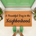 Neighborhood welcome doormat with natural coir fiber texture, featuring black script text that reads A Beautiful Day in the Neighborhood, placed on a front porch with green shoes visible for a cozy home entryway decor look.