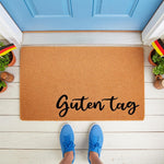 Guten Tag coir doormat featuring a natural fiber welcome mat design with black script text, placed on a white porch with blue door, German flag accents, and blue sneakers, perfect for a front door entrance or housewarming decor.