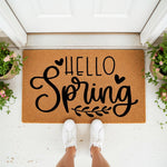 Hello Spring doormat featuring black script text and hearts on a natural coir fiber welcome mat, placed at a front door entrance with potted flowers, perfect for seasonal porch decor and outdoor entryway floor mat styling.