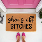 Funny doormat with the text shoes off bitches in black script and bold lettering, featuring a natural coir fiber welcome mat placed on a porch in front of a bright pink door with a person wearing pink shoes standing on the mat.