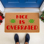Funny Christmas doormat with the text Nice Is Overrated in red letters and green snowflakes, a natural coir fiber welcome mat for holiday home decor, placed on a front porch with pine cones and holly for a festive seasonal