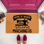 Sewing machine welcome doormat for crafters, featuring a natural coir fiber mat with black text that reads Home Is Where The Sewing Machine Is, placed on a porch floor in front of a magenta door with sewing supplies nearby.