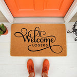 Funny Welcome Losers doormat featuring bold black script text on a natural coir fiber mat, placed on a white porch in front of an orange door with a person wearing orange sneakers standing in front of this sarcastic outdoor floor