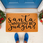 Funny Christmas doormat with the text Santa why you be judgin, a natural coir fiber welcome mat for holiday home decor placed on a porch with festive greenery and red berries, perfect for a humorous seasonal front door entrance.