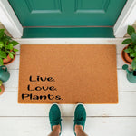 Live Love Plants coir doormat for front porch entry, featuring a natural fiber welcome mat with black text on a textured brown surface, placed on a white deck with green potted plants and a person wearing green sneakers standing