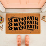 Sewciopath funny sewing doormat for crafters, featuring a wavy black text design on a natural coir welcome mat, perfect for a craft room entrance or front porch decor for anyone who loves quilting, embroidery, and sewing projects.