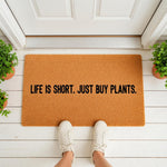 Life is short just buy plants doormat featuring a funny quote on a natural coir fiber welcome mat, placed on a white porch floor with potted greenery, perfect for a plant lover home decor entrance or a unique housewarming front