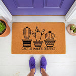 Cactus Makes Perfect doormat featuring three potted succulent illustrations on a natural coir welcome mat, placed on a white porch in front of a purple door with a person wearing purple sneakers standing on the rug for a fun