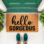 Hello Gorgeous coir doormat featuring bold black typography on a natural fiber welcome mat, placed on a front porch with green plants and a dark green door, creating a stylish entryway rug for home decor and housewarming needs.