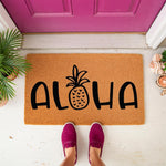 Aloha pineapple doormat for front porch entry, featuring a natural coir fiber welcome mat with black tropical lettering and a pineapple icon, placed on a white floor in front of a bright pink door for a summer home decor look.