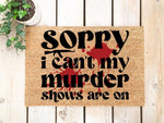 Funny murder show doormat with bold black text and red blood splatter design on natural coir fiber, perfect for true crime fans who want a humorous welcome mat for their front porch or entryway decor on a light wood floor