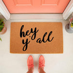 Hey Y'all doormat featuring black script text on a natural coir fiber welcome mat, placed on a front porch with a coral door and matching sneakers, creating a stylish entryway decor piece for your home or apartment front door.