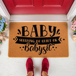 Baby Sleeping Be Quiet Or Babysit doormat, a funny natural coir fiber welcome mat for new parents featuring bold black text on a tan background, placed on a porch in front of a red door with flowers and a teddy bear nearby for