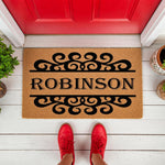 Personalized family name coir doormat featuring elegant black scrollwork design on a natural fiber welcome mat, placed on a white porch in front of a red door for a stylish home entrance decor update that adds curb appeal.