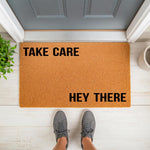 Take Care Hey There coir doormat for front porch entry, featuring a natural fiber welcome mat with bold black text on a textured brown surface, placed on a white floor in front of a gray door for a modern home decor entryway look.