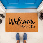 Funny Welcome Fuckers doormat made of natural coir fiber, featuring bold black text on a tan background, placed on a porch in front of a blue door with a middle finger statue nearby for a rude, sarcastic, and humorous home