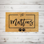 Personalized family name coir doormat with paw prints, featuring a custom last name design on a natural fiber welcome mat for pet lovers, displayed on a rustic white wood background for a stylish front porch entrance decor look.