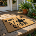 Football season doormat with Pardon the Noise text and a black football graphic, this natural coir fiber welcome mat is a funny sports decor piece for your front porch entrance during the fall game day season for any football fan