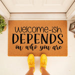 Funny welcome-ish doormat with black text on a natural coir fiber mat, placed on a front porch with yellow sneakers visible, making a humorous entryway rug for home decor that expresses a sarcastic personality to all your house