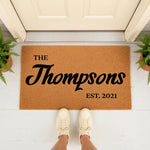 Personalized family name doormat featuring custom text on a natural coir fiber welcome mat, perfect for a front porch entryway, housewarming, or wedding decor, shown on a white floor with potted ferns and a person wearing casual