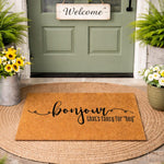 Bonjour that is fancy for hey coir doormat, a natural fiber welcome mat for front porch decor featuring black script text on a durable outdoor rug, placed on a round jute mat in front of a green door with potted flowers and