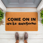 Funny welcome doormat with Come On In We Are Awesome text on a natural coir fiber mat, placed on a white porch floor with potted plants, perfect for a humorous front door entrance or a unique housewarming gift for new homeowners.