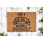 Funny doormat with the text Do I look like a people person printed in black on a natural coir fiber welcome mat, displayed on a rustic white wood background for a humorous front porch decor look that greets guests with