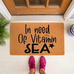 In need of Vitamin Sea doormat, a natural coir fiber welcome mat for front porch decor featuring black text and a star icon, placed on a white floor in front of a wood door with a person wearing pink sneakers standing on it.