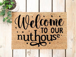 Welcome to our nuthouse coir doormat with black script lettering, a funny front porch welcome mat made of natural fiber, perfect for a humorous home entrance decor piece placed on a light wood deck with a small potted green plant.