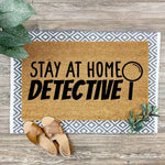 Stay At Home Detective doormat with a magnifying glass icon, featuring a natural coir fiber texture on a gray and white patterned rug base, perfect for a funny front porch welcome mat or a unique housewarming entry decor piece.