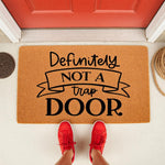 Funny Definitely Not A Trap Door doormat featuring bold black text on a natural coir fiber welcome mat, placed on a porch in front of a red door with a person wearing red sneakers standing on the mat for a humorous home entrance