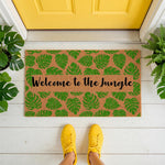 Tropical leaf welcome doormat with Welcome to the Jungle text, featuring a natural coir fiber material and green monstera leaf pattern, placed on a white porch in front of a bright yellow door for a stylish home entryway decor