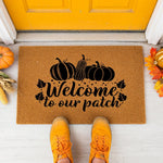 Fall pumpkin welcome doormat featuring Welcome to our patch text and black pumpkin graphics on a natural coir fiber mat, displayed on a porch with autumn leaves and corn decor for seasonal front door entry home entryway floor