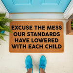 Funny doormat with the text Excuse the Mess Our Standards Have Lowered With Each Child, a natural coir fiber welcome mat for front porch decor that makes a humorous housewarming gift for parents and families with kids at home.