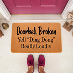 Funny Doorbell Broken Yell Ding Dong Really Loudly doormat, a natural coir welcome mat for front porch decor that features humorous text, placed on a white floor in front of a magenta door with a person wearing maroon sneakers