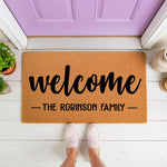 Personalized family name welcome doormat featuring a natural coir fiber texture and bold black script text, placed on a white porch in front of a purple door as a custom housewarming entry rug for a new home or front door decor.