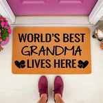 World's Best Grandma Lives Here doormat featuring bold black text and heart icons on a natural coir fiber welcome mat, placed on a front porch entryway to celebrate a beloved grandmother with a stylish and durable home decor