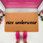 Funny nice underwear doormat made of natural coir fiber, featuring bold black text on a tan welcome mat surface, placed on a porch in front of a bright pink door for a humorous home decor entrance statement that guests will love.