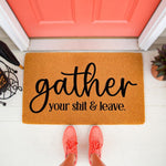 Funny doormat with rude text gather your shit and leave printed in black script on a natural fiber welcome mat, placed on a porch in front of a coral door with a person wearing coral sneakers standing on the entryway floor mat.