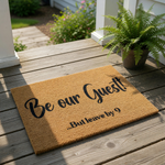 Funny Be Our Guest But Leave By 9 doormat featuring bold black text on a natural coir fiber welcome mat, displayed on a front porch wood deck with a green fern plant nearby for a humorous and inviting home entryway decor look.