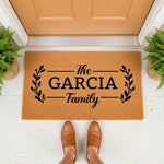Personalized family name doormat featuring a natural coir fiber texture with black leaf branch accents, serving as a stylish front porch welcome mat for a home entrance, displayed on a white floor with potted ferns and brown
