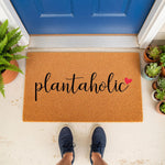 Plantaholic coir doormat for plant lovers, featuring a natural fiber welcome mat with black script text and a small red heart, displayed on a front porch with potted succulents and a fern by a blue door for a stylish home