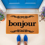 Bonjour coir doormat with black script text and decorative flourishes, placed on a porch in front of a blue door, making a stylish welcome mat for your home entrance or front door decor for a modern house entryway.