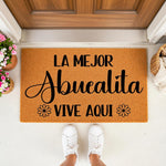 La Mejor Abuelita Vive Aqui doormat, a natural coir welcome mat for a grandmother featuring black script text and floral accents, placed on a front porch entryway with potted flowers and a basket, perfect for a home entrance