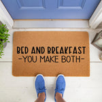 Funny Bed and Breakfast You Make Both doormat featuring bold black text on a natural coir fiber welcome mat, placed on a white porch in front of a blue door for a humorous home decor entryway statement that guests will surely