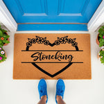 Personalized family name doormat featuring a black floral heart design on natural coir fiber, perfect as a custom housewarming welcome mat for your front porch entrance, shown with blue sneakers on a white floor by a blue door.