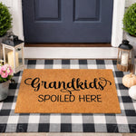 Grandkids Spoiled Here doormat, a natural fiber welcome mat for grandparents featuring black script text, displayed on a black and white buffalo plaid rug at a front door entrance for a cozy, farmhouse porch decor look.